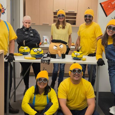 Group of people in an office dressed as Minions from Despicable Me, most wearing yellow shirts, blue overalls, and hats, with one individual dressed as Gru, standing and sitting around a table decorated with Minion-themed pumpkins