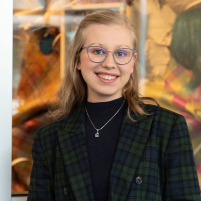 Person wearing a dark plaid blazer over a black top with a silver necklace, standing in front of a colorful patterned textile display