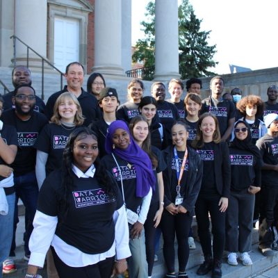 Group of people wearing matching black ‘Data Warriors’ T-shirts standing together on outdoor steps in front of a building with columns.