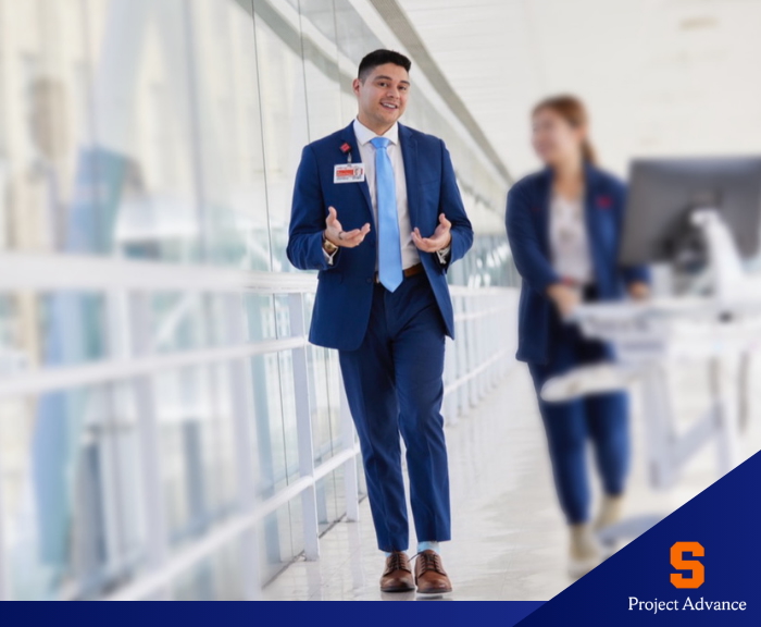 Two professionals in blue suits walking through a bright, modern hallway with glass walls, one speaking while the other pushes a mobile workstation. A logo in the bottom corner reads “S Project Advance.