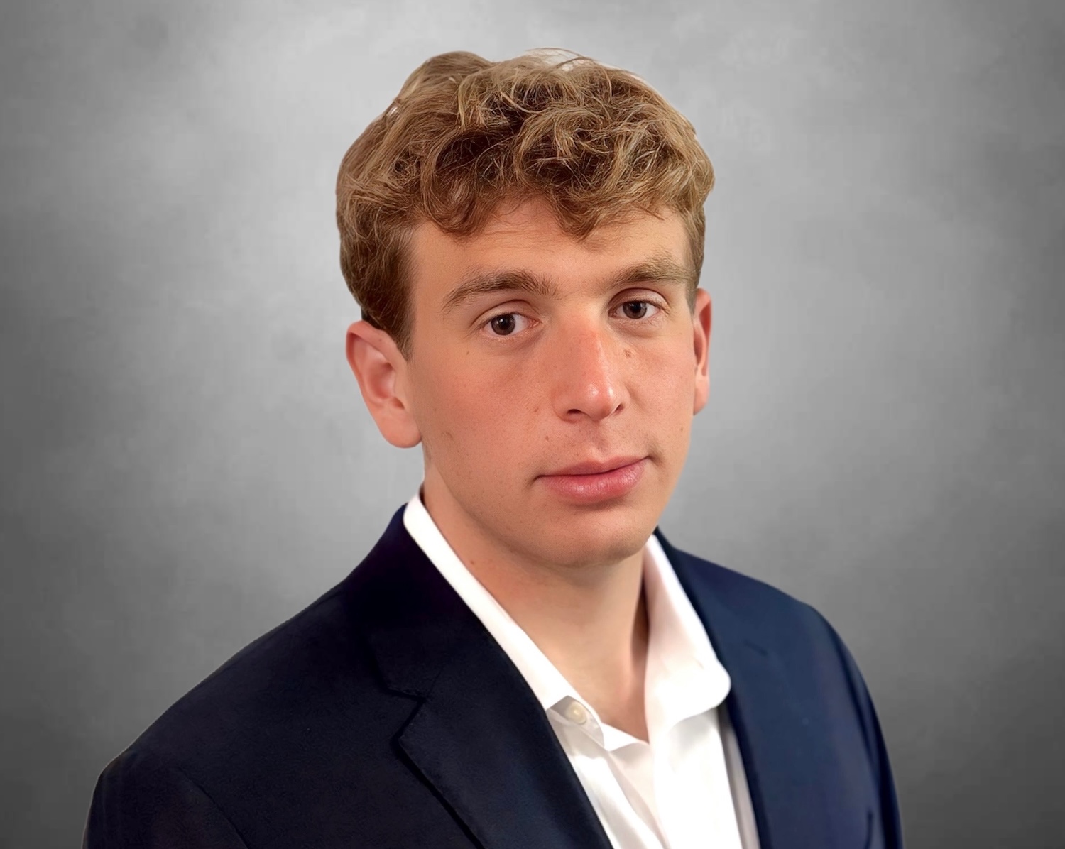 Person in a dark suit jacket and white dress shirt posed against a plain gray background.