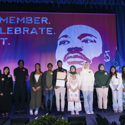 A group of people standing on a stage in front of a large banner featuring an illustration of Martin Luther King Jr. and the words “REMEMBER. CELEBRATE. ACT.”