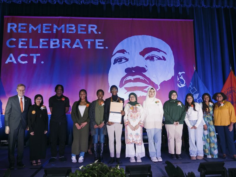 A group of people standing on a stage in front of a large banner featuring an illustration of Martin Luther King Jr. and the words “REMEMBER. CELEBRATE. ACT.”