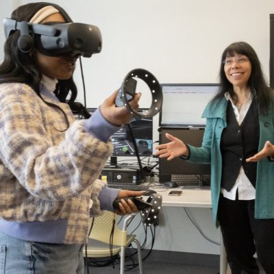 Professor T. Makana Chock observes a student wearing a virtual reality headset and holding VR controllers in the Extended Reality Lab.