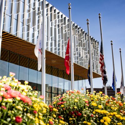 Flags displayed in front of a modern glass building with colorful flowers in the foreground.
