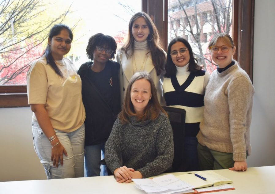 Six women pose together in an office or classroom setting with large windows showing bare trees and buildings outside. Five women stand in the back row, while one woman sits at a table in the front.