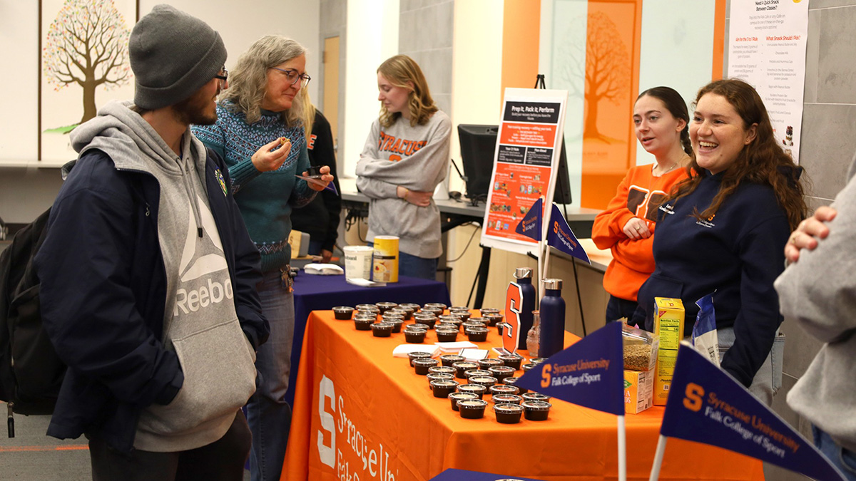 Campus event featuring a nutrition and wellness table displaying numerous small sample cups, informational pamphlets, and mini flags. A large orange tablecloth with the Syracuse University Falk College of Sport logo covers the table, and a poster board with health-related information stands behind it. Several people are gathered around, engaging in conversation.