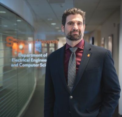A person wearing a dark suit jacket, maroon dress shirt, patterned tie, and a lapel pin stands in a modern hallway. Behind them is a glass wall with the text “Syracuse University Department of Electrical Engineering and Computer Science” visible.