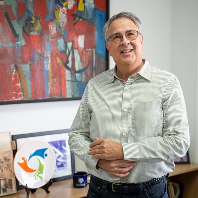 A man wearing glasses, a green shirt and jeans in his office. There is colorful artwork in the background