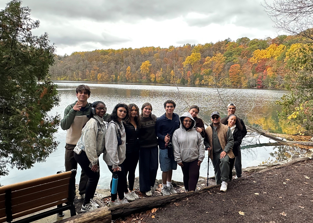 Group of people standing together near a lakeside with autumn-colored trees in the background. A wooden bench and fallen log are in the foreground, and the lake reflects the colorful foliage under a cloudy sky
