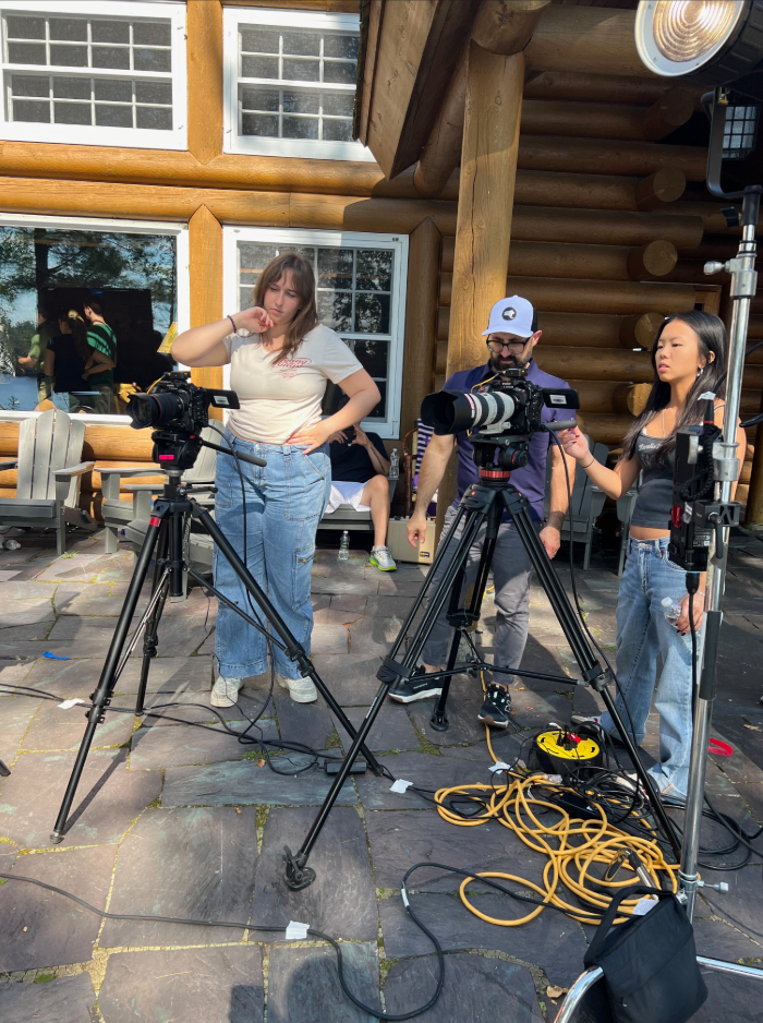 Two professional video cameras on tripods set up on a stone patio outside a log cabin, with lighting equipment and coiled cables on the ground.
