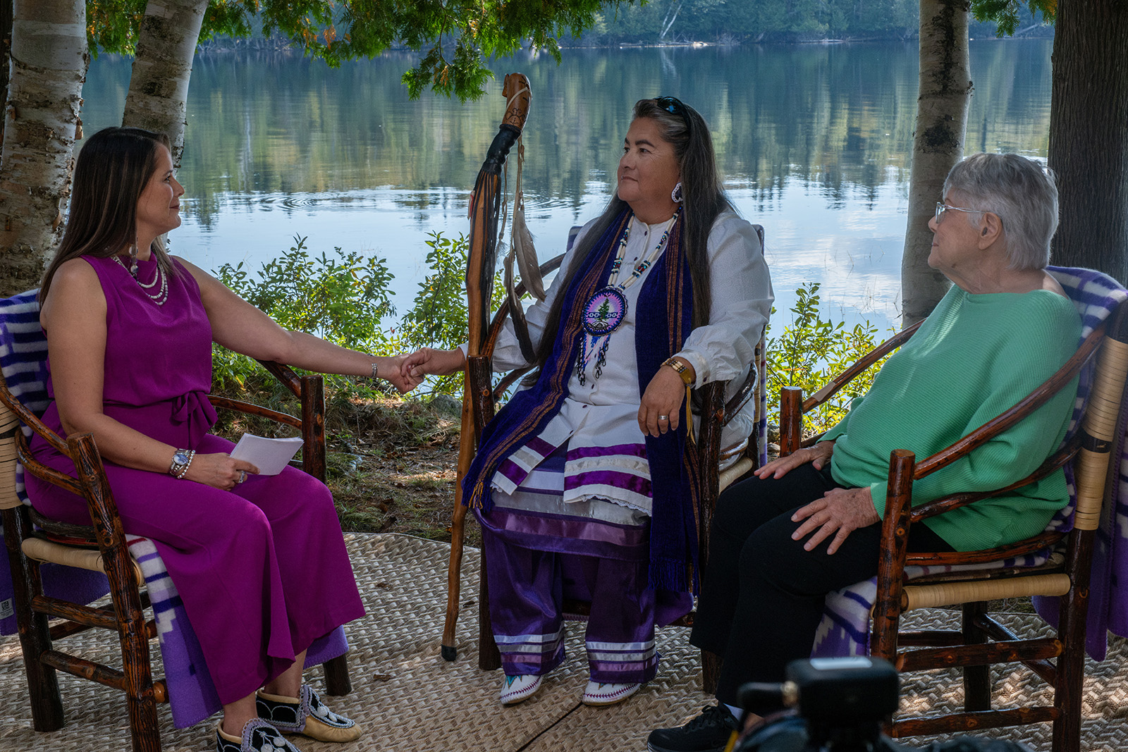 Three people seated on wooden chairs with woven blankets, engaged in conversation near a calm lake, surrounded by trees and natural greenery