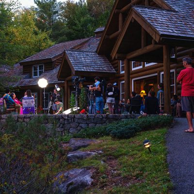 Outdoor film crew setting up cameras and lights on a stone patio in front of a large log cabin, with several people gathered around watching the scene
