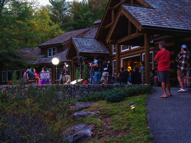 Outdoor film crew setting up cameras and lights on a stone patio in front of a large log cabin, with several people gathered around watching the scene