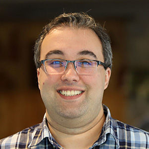 Dark-haired man, smiling and wearing glasses, wearing a plaid shirt