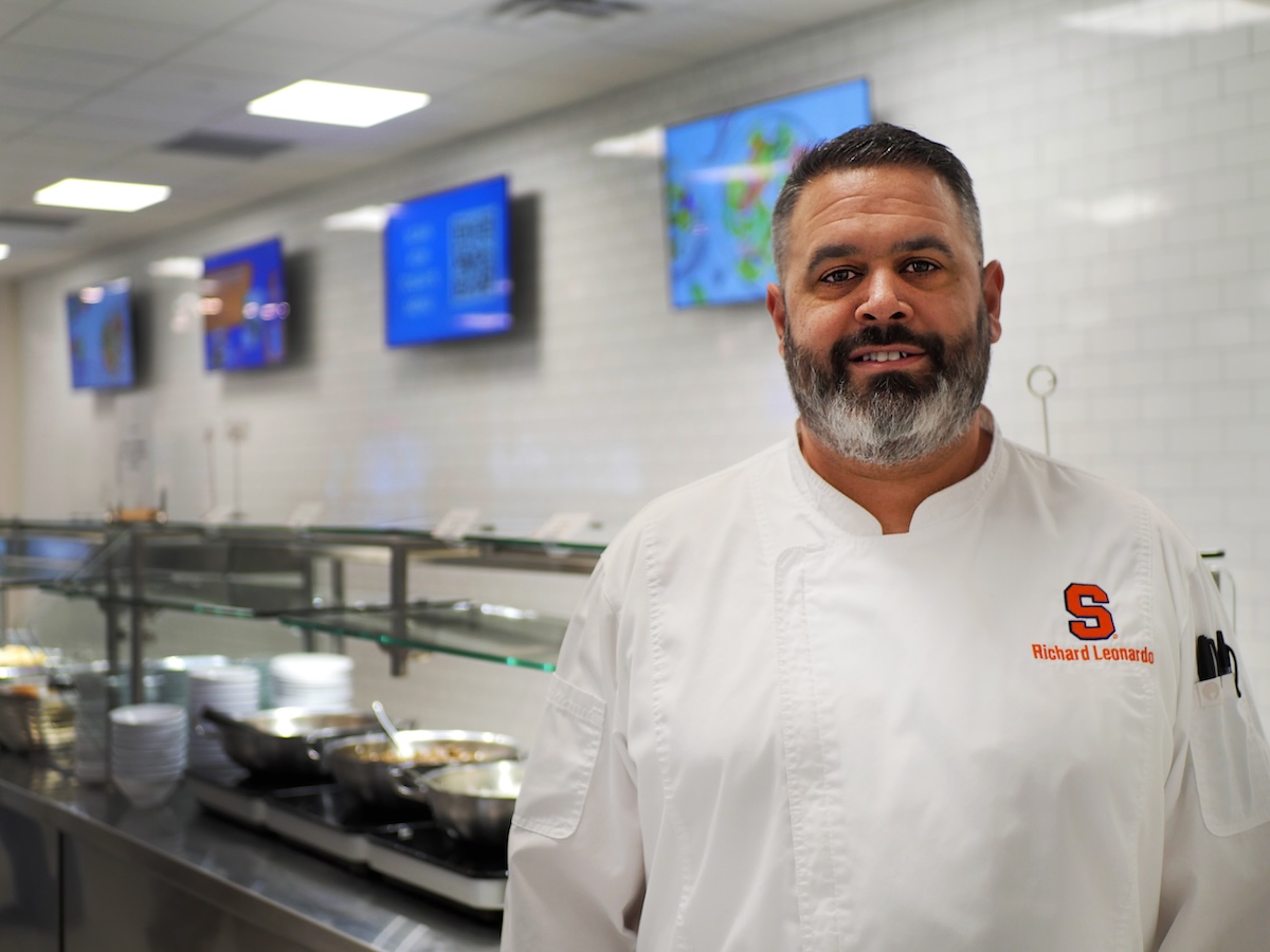 Chef in a white uniform standing in a cafeteria-style kitchen with stainless steel counters, serving pans, and plates.