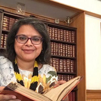 Woman with glasses, smiling and wearing a colorful dress and scarf, holds an open book. A bookshelf filled with books is behind her.