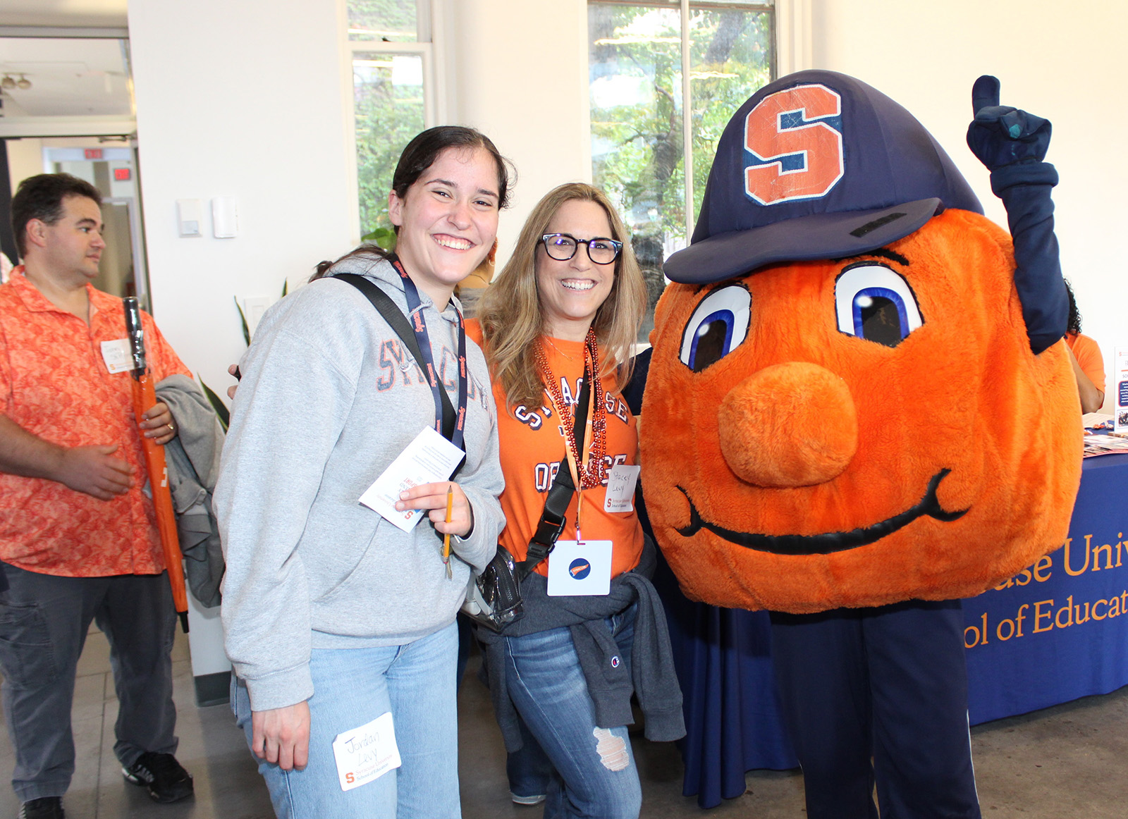 Three people posing with Otto the Orange mascot wearing a blue cap and jersey with an ‘S’ logo at a Syracuse University event