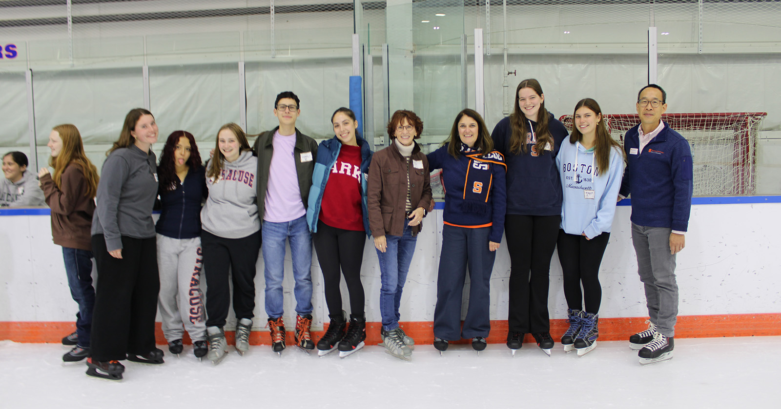 Group of people standing together on an indoor ice rink, wearing casual winter clothing and ice skates, with hockey boards and a goal net in the background