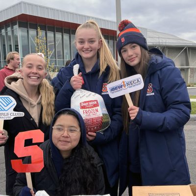 Four student-athletes pose with a frozen turkey and promotional signs for the Boys & Girls Clubs of Syracuse outside a campus building.