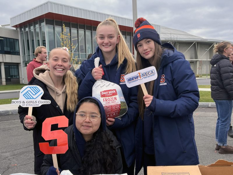 Four student-athletes pose with a frozen turkey and promotional signs for the Boys & Girls Clubs of Syracuse outside a campus building.