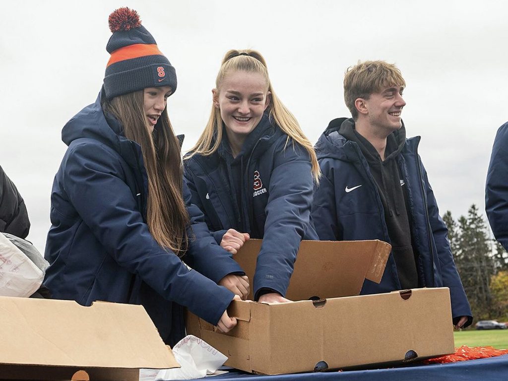 Three students in Syracuse athletic gear smile while packing cardboard boxes at an outdoor service event.