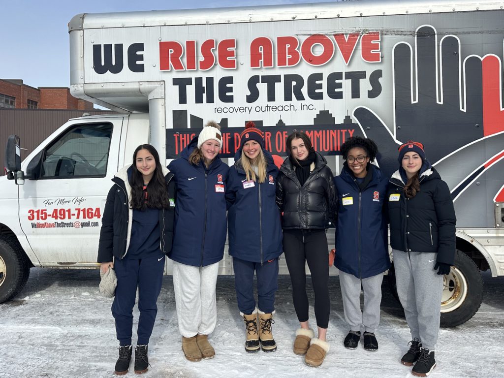 Six student-athletes in Syracuse apparel stand in front of a "We Rise Above the Streets" recovery outreach van in winter conditions.