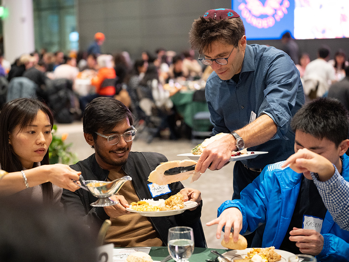 People are seated around a table with food, a man is standing and serving turkey