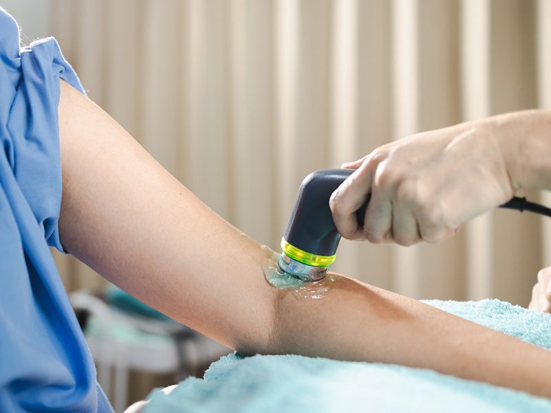 An ultrasound is performed on the left arm of a patient in a medical gown.