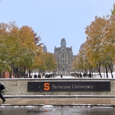 Syracuse University campus in winter with light snow, colorful autumn trees, and the historic Hall of Languages building in the background.
