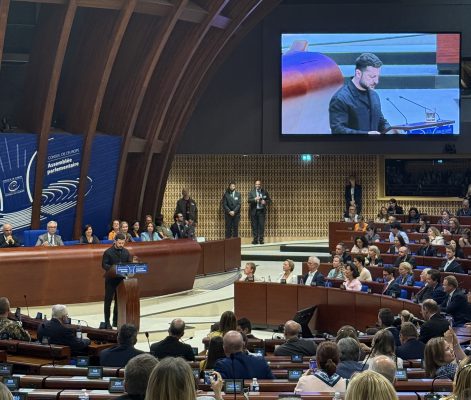 An individual giving a speech at the Council of Europe. The chamber is filled with seated attendees and features a large screen showing the speaker.