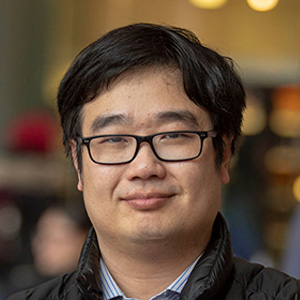 Dark-haired man with glasses, smiling, wearing a blur shirt and dark jacket