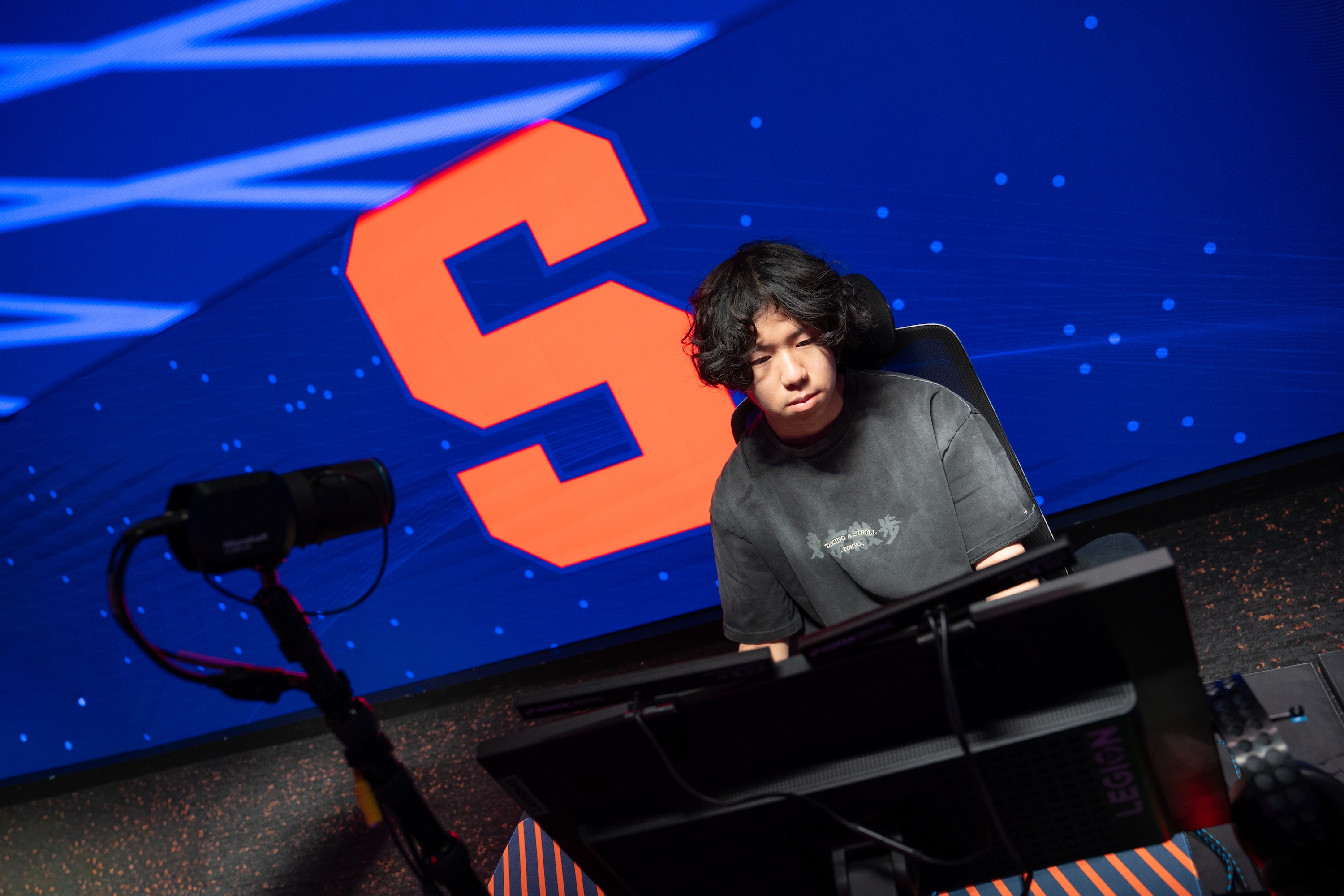 Person seated at a gaming station with monitors and a camera setup, in front of a large screen displaying an orange “S” on a blue background