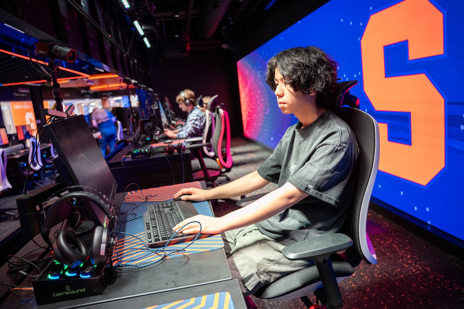 Person seated at a gaming station with keyboard and monitor, wearing a headset, in a row of players inside an esports arena with a large orange “S” on a blue background wall.