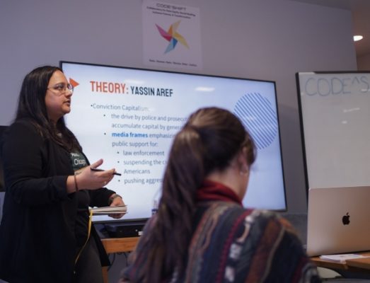 A young person wearing glasses and a black shirt presents to an audience, gesturing toward a screen displaying a slide titled "THEORY: YASSIN AREF" with bullet points about "Conviction Capitalism" and media frames. An audience member with long dark hair is visible in the foreground.
