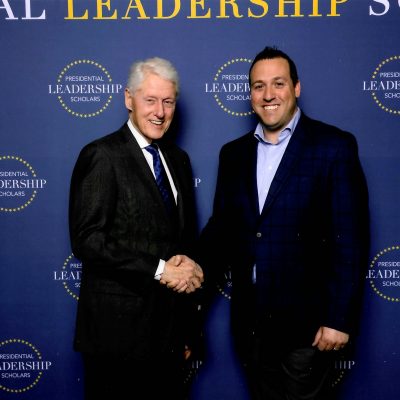 Two individuals in formal attire shaking hands in front of a blue backdrop with repeated “Presidential Leadership Scholars” logos and text.