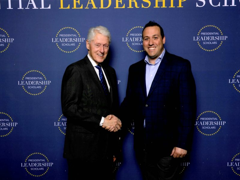 Two individuals in formal attire shaking hands in front of a blue backdrop with repeated “Presidential Leadership Scholars” logos and text.