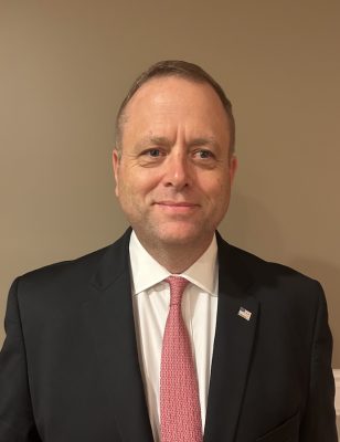 Person wearing a dark suit jacket with a white dress shirt and a pink patterned tie, featuring a small U.S. flag lapel pin