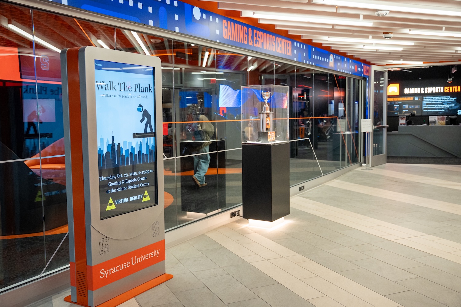 Digital kiosk near a glass wall showcasing a trophy inside the Syracuse University Gaming & Esports Center.