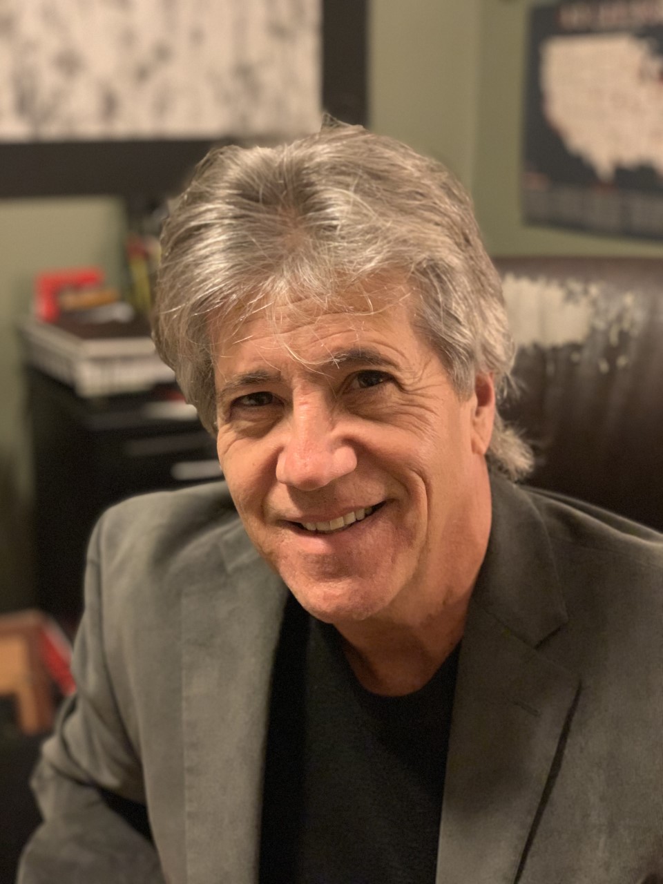 Person wearing a gray blazer over a black shirt, seated indoors with a dark desk and shelves in the background. 