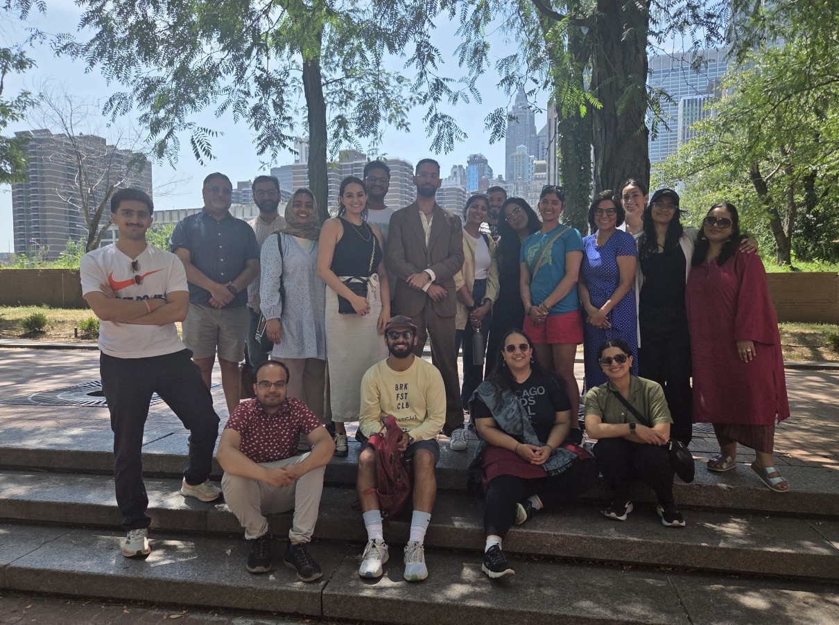 A diverse group of approximately 20 people poses for a photo in an urban park, with some standing and four sitting on stone steps in the front row. The group is dressed in a mix of casual and business casual attire, including traditional South Asian clothing. Behind them, leafy trees frame a city skyline.