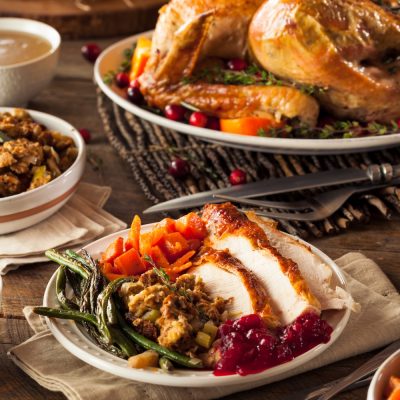 Plate of sliced turkey with stuffing, green beans, carrots, and cranberry sauce, with a roasted turkey and side dishes in the background.