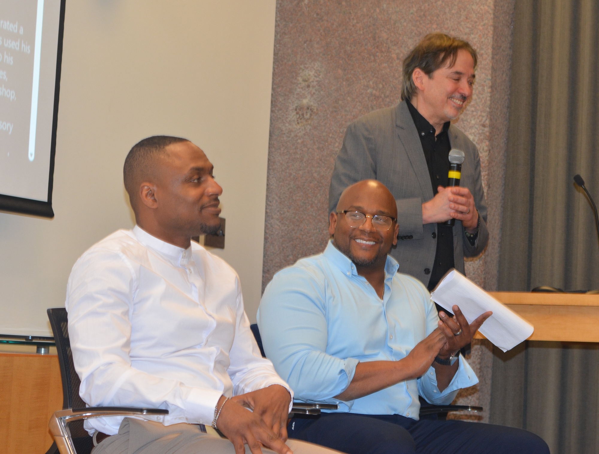 Two people seated with papers and a phone, while another person stands at a podium holding a microphone in a conference or presentation setting.