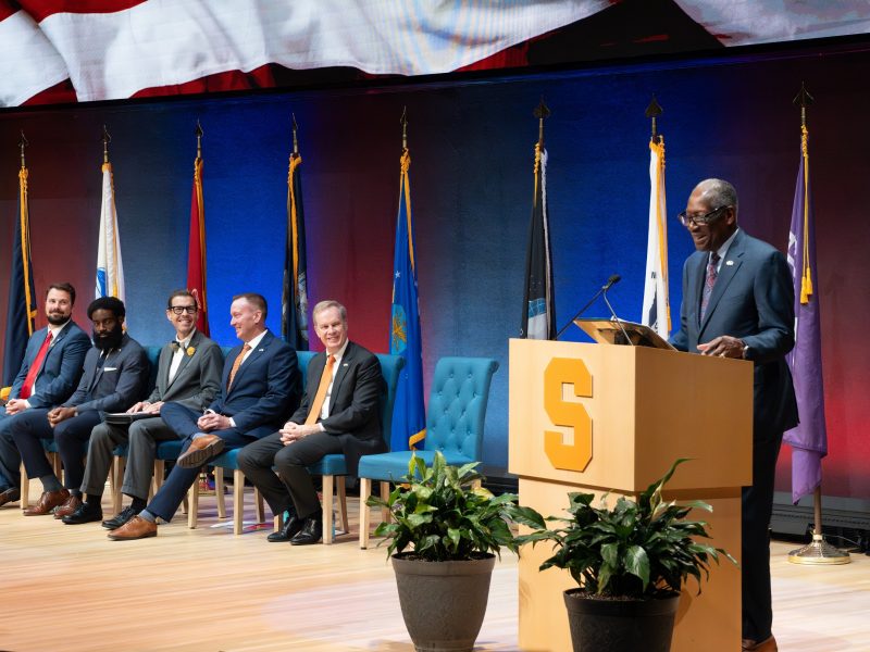 person speaking at podium with five people seated to his right in front of several flags