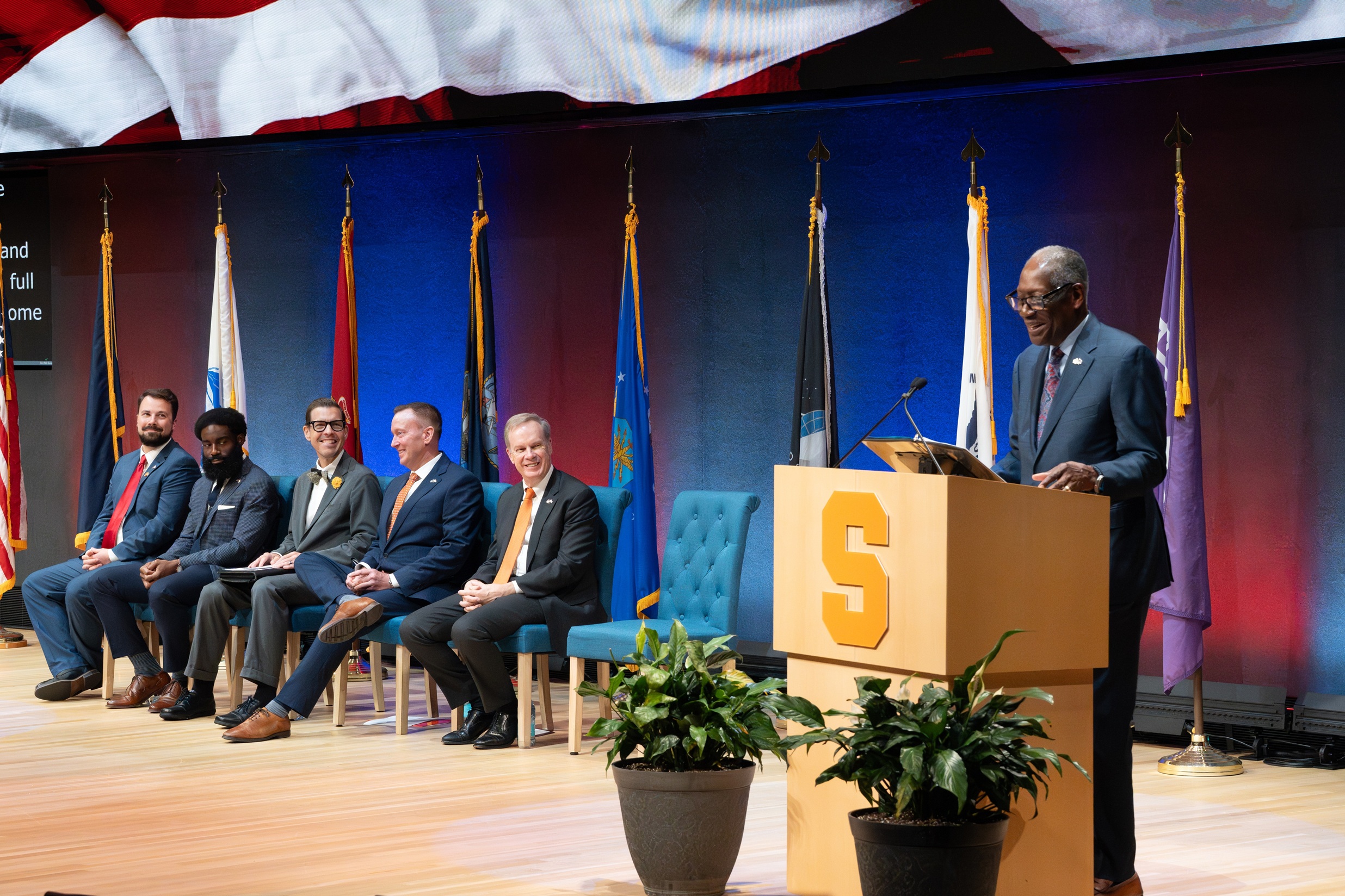 person speaking at podium with five people seated to his right in front of several flags