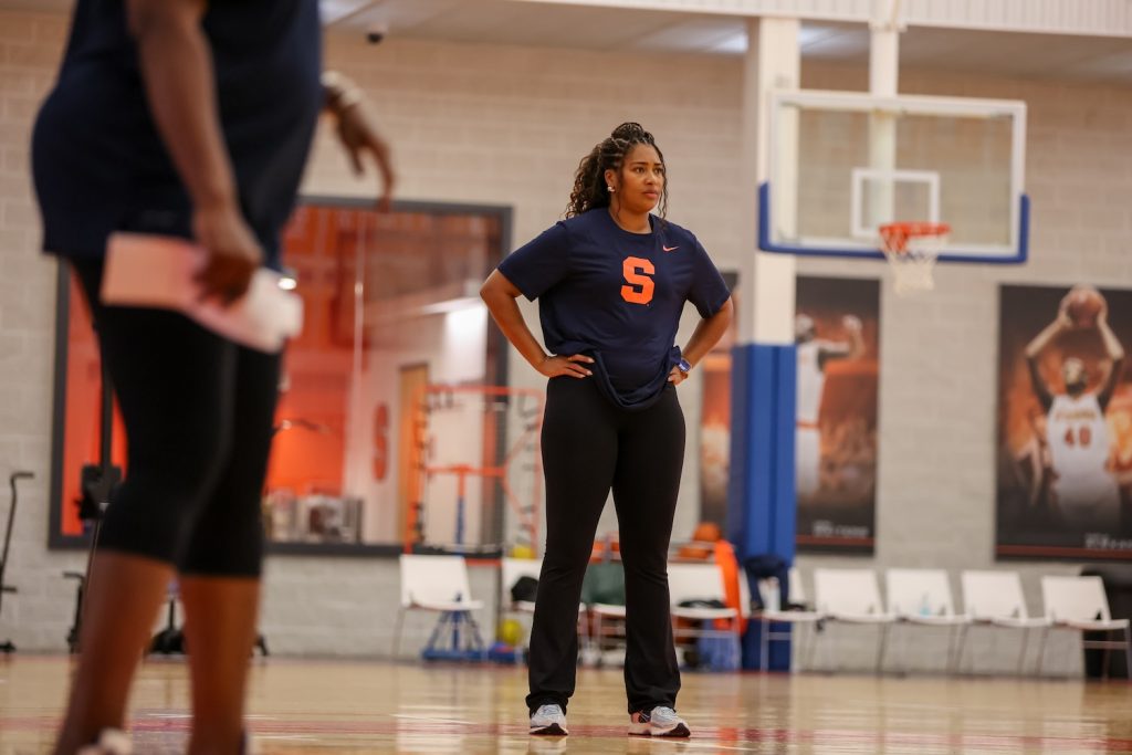 Individual standing on a basketball court wearing a navy blue shirt with a large orange “S” logo and black pants, hands on hips; another person partially visible in the foreground holding papers