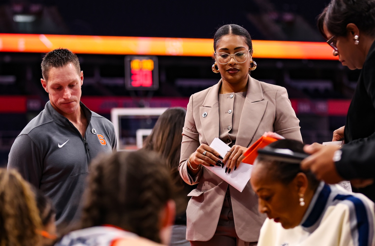 Two individuals standing courtside during a basketball game; one is in a tan suit holding papers, and the other in a dark athletic jacket. Players are seated in the foreground