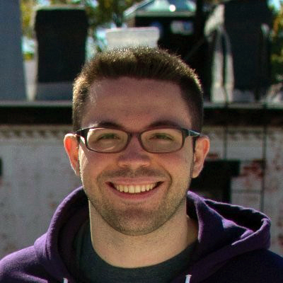 Professional headshot of a smiling man with glasses and short hair wearing a purple hoodie, photographed outdoors with natural lighting.
