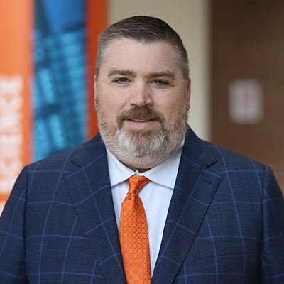 Professional headshot of Syracuse University administrator in navy windowpane suit and orange tie against blurred campus background.