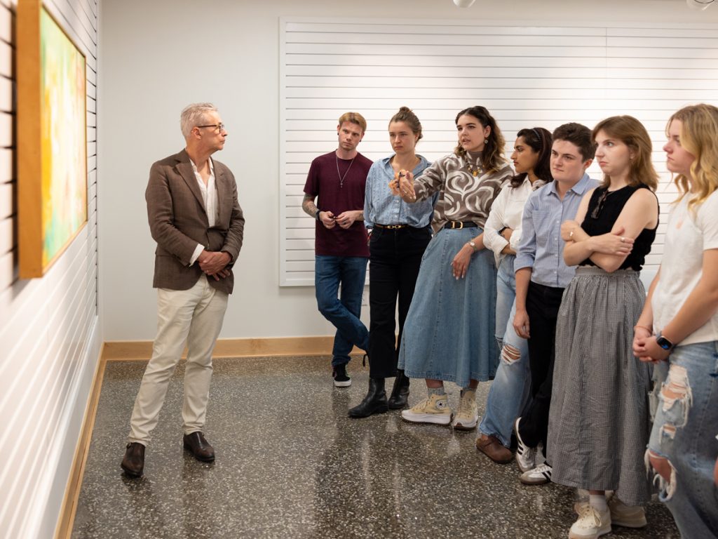 A person in a brown blazer gestures toward artwork on a gallery wall while teaching a group of students who stand listening attentively in a white-walled exhibition space.